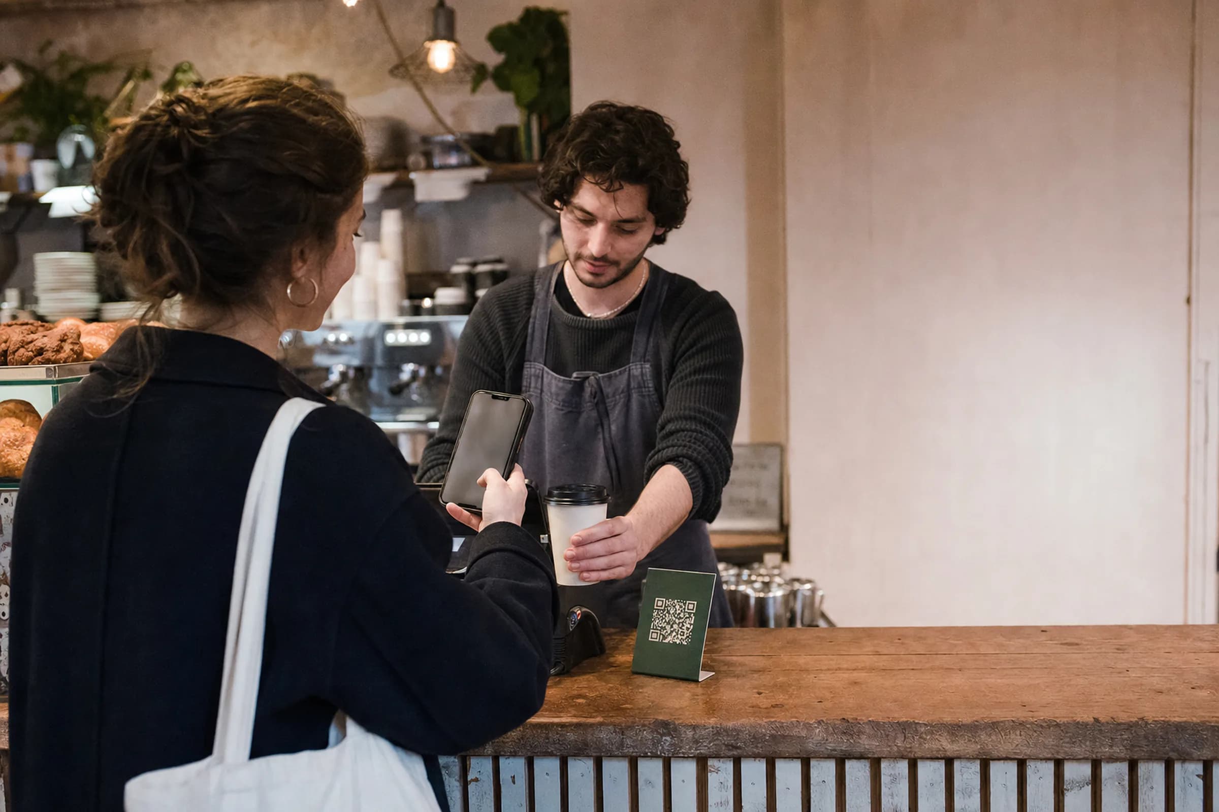 A customer holding their phone up to a small Coco Regulars stand on a café counter while the barista serves them